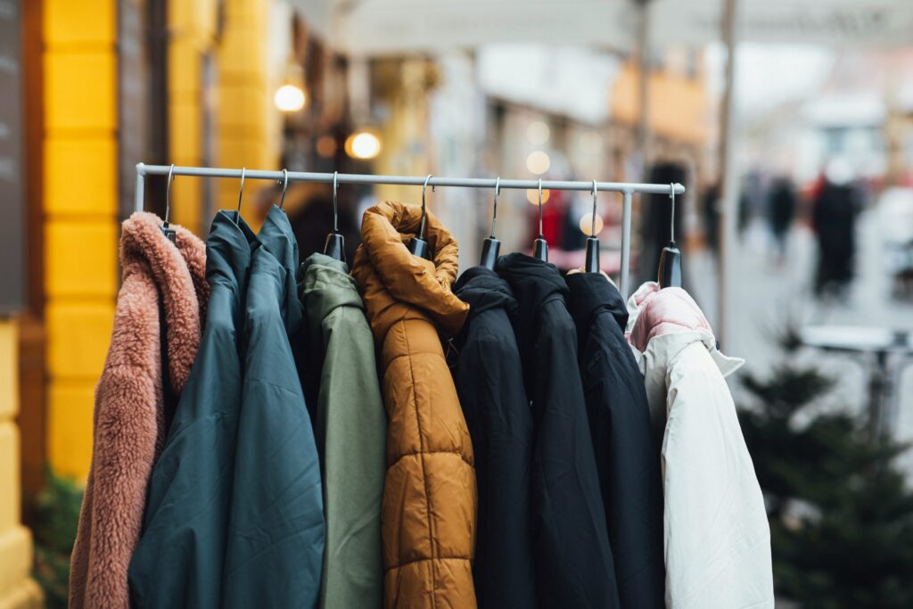 A variety of colorful and stylish jackets and coats hanging on a metal rack outdoors, perfect as fashion gifts for men. The collection includes different textures, like a sherpa jacket and puffer coats, in warm colors such as burnt orange, black, green, and cream, suggesting a versatile selection of  men's winter outerwear.