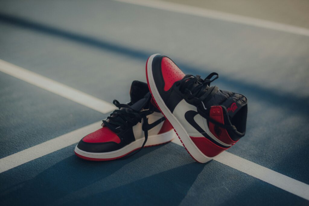 A pair of high-top stylish sneakers in a classic black, white, and red colorway (resembling a Jordan 1), resting on a blue track or court. These trendy yet timeless athletic shoes are a perfect fashion gift idea for men who appreciate comfortable, stylish and high-quality footwear.