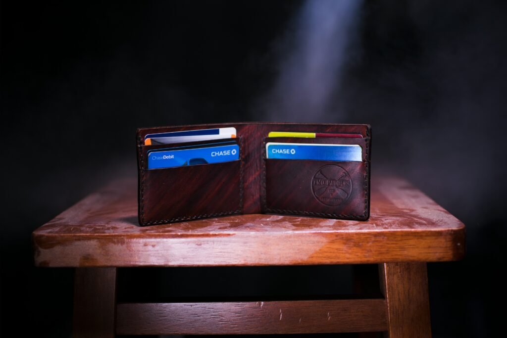 An open, dark brown or burgundy personalized leather wallet sitting on a small wooden table, partially illuminated by a beam of light. The wallet is folded open to show several credit cards and highlights a quality men's accessory and a thoughtful gift idea that combines style and sentimentality.