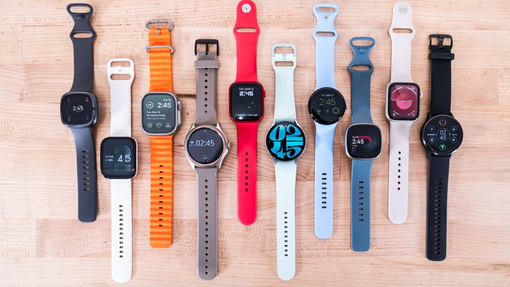 A flat lay of nine diverse men's and unisex smartwatches on a wooden surface, ranging in colors like black, white, orange, grey, red, and blue. Smartwatch brands visible include Fitbit and Samsung Galaxy Watch, representing modern gift ideas for men interested in tech, fitness, and health tracking.