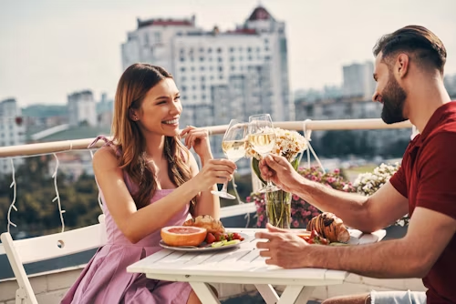 Romantic dinner date gift idea: A happy couple toasting wine on a balcony, illustrating a perfect "take her for dinner" experience for the "Best Gift Ideas for Women" post.