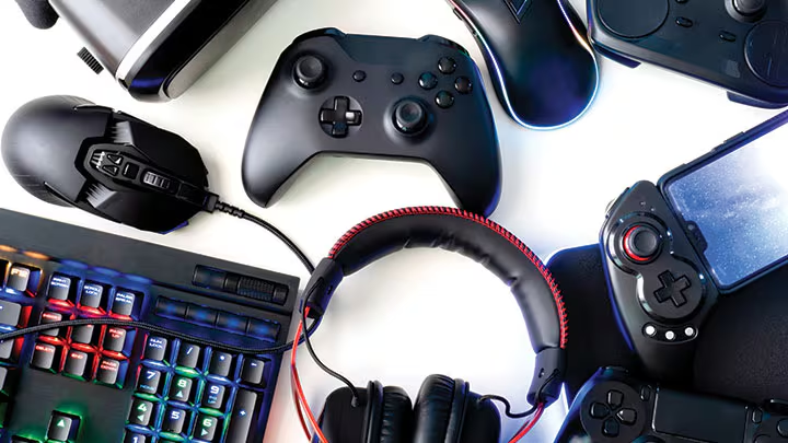 A flat lay of essential gaming accessories arranged on a white surface, including a black Xbox controller, a high-performance **gaming mouse**, a vibrant **RGB keyboard**, a padded over-ear **gaming headset**, and a handheld console. This collection represents great tech gift ideas for men and gamers who want to upgrade their dream setup.
