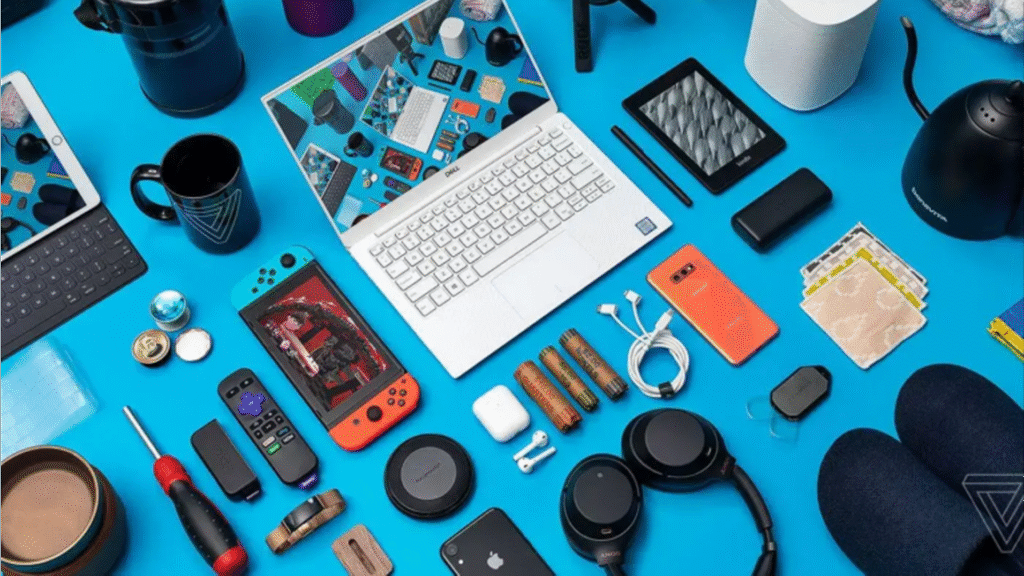 A vibrant flat lay of essential tech gadgets and  accessories laid out on a bright blue surface, centered around a laptop computer. The collection of must-have gifts for men** includes a smartphone, wireless headphones, a portable speaker, a Nintendo Switch, tablet, power banks, mugs, and other small electronic devices, representing a modern collection of cool technology gifts.