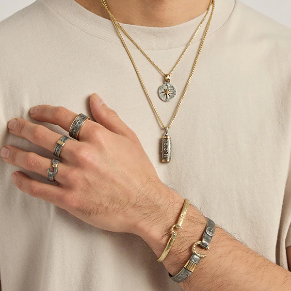 A close-up of a man's arm and chest showcasing several pieces of personalized jewelry, including layering gold necklaces with engraved or initial pendants, **rings**, and matching **bracelets**. This subtle and stylish accessory set is a thoughtful and memorable gift idea for men who appreciate sentimental or custom items.