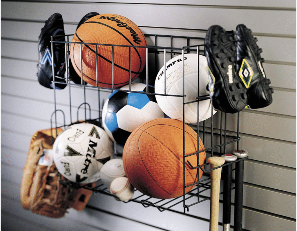 A wire mesh display bin filled with various pieces of sports gear and athletic equipment, including basketballs, soccer balls, volleyballs, and two pairs of black cleats hanging on the rack. This collection is a great selection of gifts for men and competitive guys interested in team sports and athletic activities.