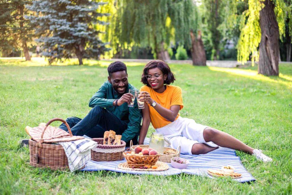 Happy Black couple enjoying a romantic summer picnic date in a sunny park, clinking glasses and smiling. Best outdoor date ideas for 2025, summer dates, affordable romantic activities, couple picnic, relationship with murf blog.