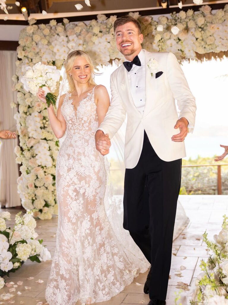 Newlyweds MrBeast and his bride walking down the aisle holding hands with flower petals falling around them.