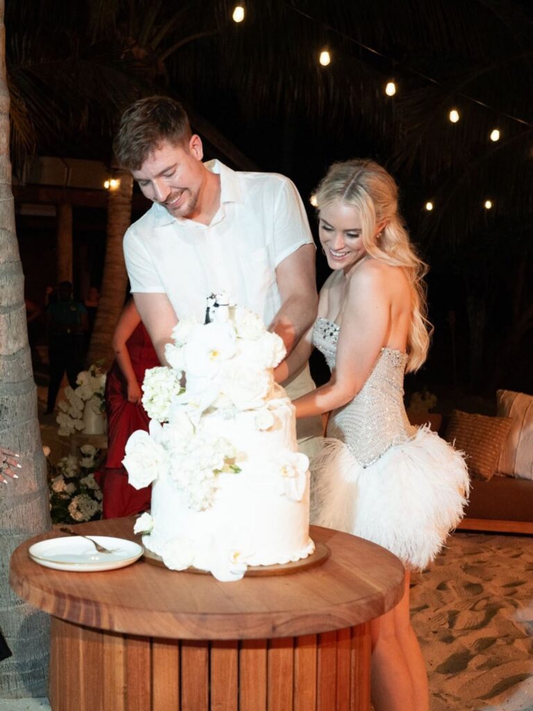 MrBeast and his bride together cutting a multi-tiered floral wedding cake under string lights outdoors.